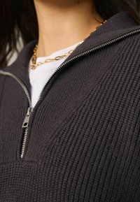 Pullover zip-up nero realizzato in tessuto a coste, con colletto alto e zip argentata, indossato sopra una maglietta bianca. Visibile una collana in oro.
