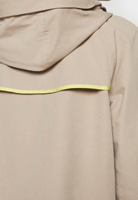 Beżowa, lekka kurtka z kapturem, z poziomym żółtym odblaskowym akcentem na plecach. Gładka tekstura, prosty design.