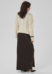 Kvinde med mørkt hår iført en cremet kabelstrik sweater, sort satin ankel-længde nederdel, sorte kitten heels og et sort armbånd, stående sidelæns.