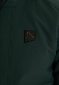 Zelena jakna z matiranjem, z kvadratno črno nalepko in oranžnim šivom na levi prednji strani.