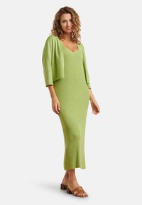 Groene gebreide midi-jurk met een V-hals en ribbelstructuur, gecombineerd met een bijpassende cropped cardigan en bruine blokhak-sandalen.