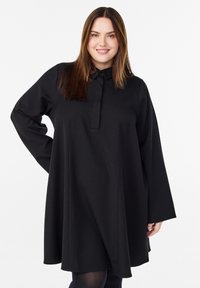 Usmívající se žena se lhavými hnědými vlasy, oblečená v volných černých dlouhých šatech s límečkem, na bílém pozadí.