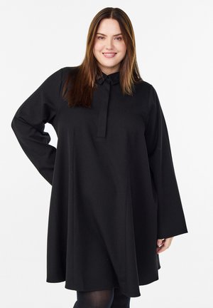 Smilende kvinde med glat brunt hår iført en løs sort langærmet kjole med krave foran en hvid baggrund.