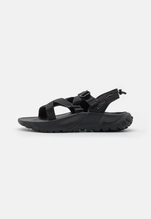 Sandale de sport noire avec semelle épaisse, brides réglables sur le pied et autour du talon, et semelle texturée pour usage extérieur.