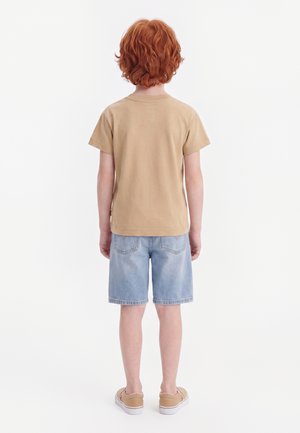 Enfant aux cheveux roux bouclés portant un t-shirt beige, un short en jean bleu clair et des baskets marron, debout de dos sur fond blanc.