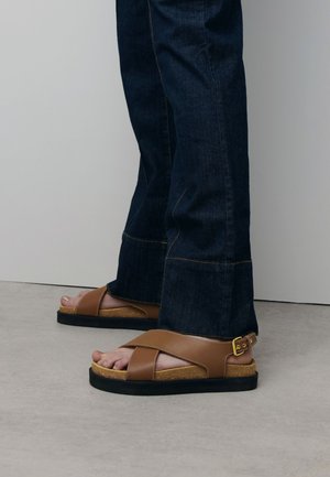 Brown leather sandals with cross-strap design, cork sole, and black rubber base, worn with dark denim jeans, showing exposed toes.