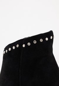 Collar di stivaletti neri in suede alla caviglia con una fila di piccoli chiodi tondi in argento lungo il bordo superiore, su sfondo bianco.