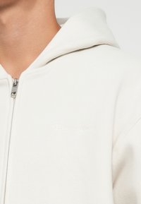 Felpa zip con cappuccio di colore crema realizzata in tessuto morbido, con un logo ricamato discreto sul petto e hardware della cerniera in argento.
