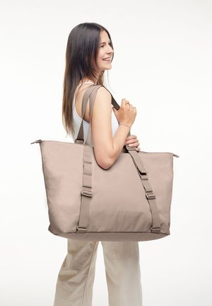 Femme souriante, portant un grand sac fourre-tout beige avec deux anses, vêtue d'un débardeur blanc et d'un pantalon clair, se tenant devant un fond blanc.