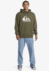 Olive green hoodie with kangaroo pocket and white logo, paired with light blue jeans and black sneakers. Soft fabric and relaxed fit.