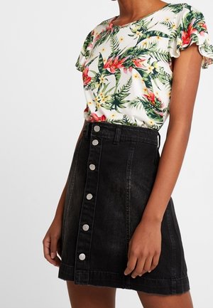 Vrouw draagt een witte korte mouwen top met bloemenprint, ingestopt in een zwarte, hooggetailleerde denimrok met zilveren knopen aan de voorkant.