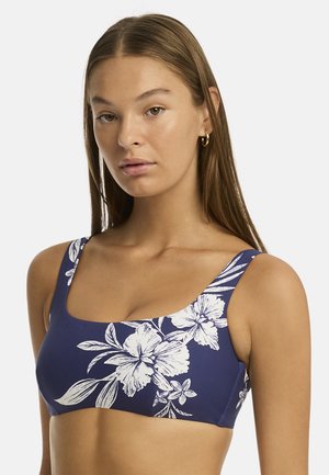 Młoda kobieta w niebieskim topie bikini z białym kwiatowym wzorem, długie proste włosy i mała złota obrączka w uchu na białym tle.