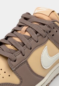 Nike-sneakers med en beige och brun färgskala, slätt syntetmaterial, runda snören och en framträdande vit logotypaccents.