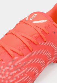 Φωτεινό κοραλλί ποδοσφαιρικό παπούτσι από λείο συνθετικό υλικό, με λεπτές λευκές ρίγες και κλείσιμο με κορδόνια, με έντονη λεπτομέρεια εμπορικού σήματος.