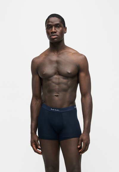 Homme musclé debout, portant un boxer bleu marine foncé avec le logo Paul Smith sur la ceinture, sur un fond uni gris clair.