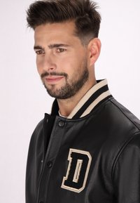 Veste varsity en cuir noir avec col strié beige et un patch en forme de "D" texturé sur la poitrine. Texture lisse et fermeture à boutons-pression.