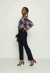 Blusa preta fluida com estampado floral vibrante, combinada com calças pretas ajustadas com laterais fendidas e sapatos pretos de salto aberto.