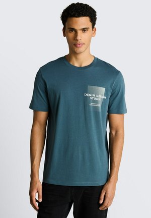 Man met krullend haar draagt een turquoise korte mouwen t-shirt met witte tekst "Denim Design Studio" op het borstzakgebied.