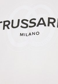 Logo Trussardi Milano en police noire en gras sur fond blanc avec un emblème circulaire discret derrière le texte.