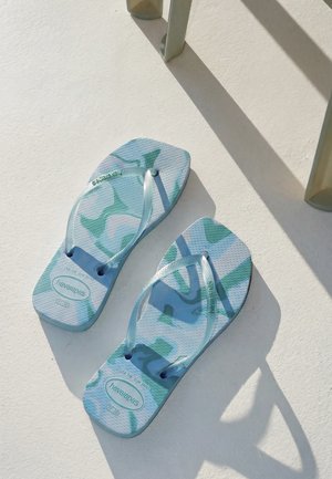 Blå og grøn mønstrede Havaianas flip-flops på en hvid overflade med nærliggende stolskygger.