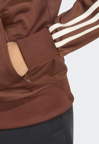 Veste marron zippée avec un tissu texturé, des poignets côtelés et trois rayures blanches le long des manches. Comprend un détail de poche zippée sur le côté.
