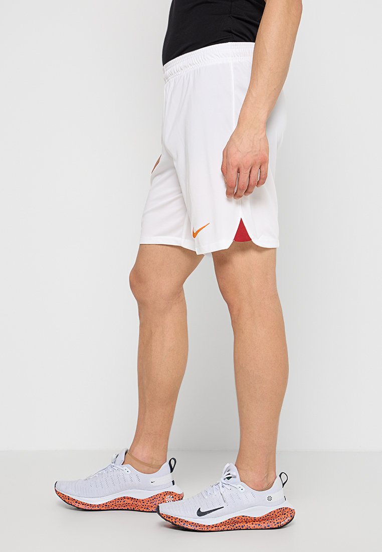 Białe sportowe szorty z pomarańczowym logo Nike, z czerwonymi akcentami po bokach. Noszone z jasnoszarymi butami z pomarańczowymi detalami.