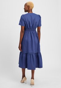 Navy blue, knee-length dress made of lightweight fabric. Features puff sleeves, smocked waist, and tiered ruffle hem. Worn with beige heels.