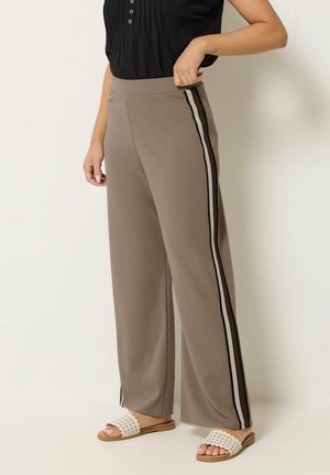 Femme portant un pantalon taupe à jambes larges avec des bandes latérales noires et blanches, un haut noir sans manches et des sandales blanches à enfiler.