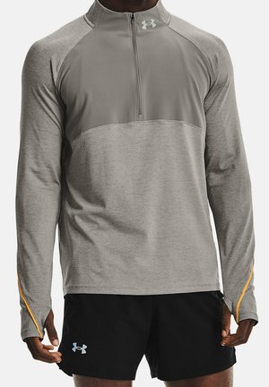 Mies, joka pukeutunut vaaleanharmaaseen neljänneksen vetoketjulliseen urheilupaitaan, jossa on peukaloaukot, sekä mustiin urheilushortseihin, joissa on logot rinnassa ja lahkeessa.