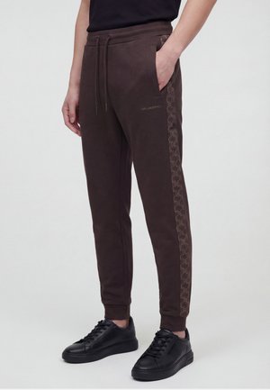 Person trägt dunkelbraune Jogginghosen mit seitlichem Musterstreifen und schwarzen Sneakers, Hand in der Tasche, steht vor hellem Hintergrund.