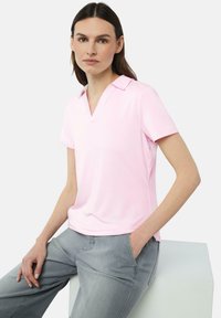 Hellrosa Poloshirt mit V-Ausschnitt und kurzen Ärmeln, aus weichem Stoff. Kombiniert mit grauen Hosen, Model sitzt auf einer weißen Fläche.