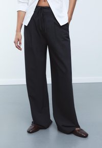 Pantalons larges à rayures noires avec une taille à cordon de serrage, dotés de plis et d'une texture lisse, associés à des mocassins marron.