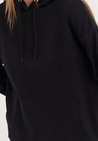 Sudadera negra con capucha y bolsillo frontal, confeccionada en un tejido suave. Cuenta con cordones ajustables y un corte holgado con puños acanalados.