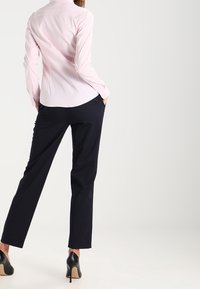 Kobieta w jasnoróżowej koszuli zapinanej na guziki, czarnych spodni z lekkiego materiału oraz czarnych szpilkach, stojąca z rękami w kieszeniach, tyłem do widza.