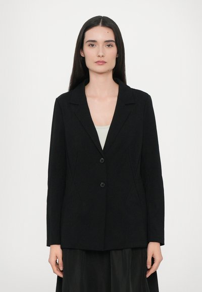 Blazer nero con colletto a punta, due bottoni neri, vestibilità su misura e texture liscia, abbinato a un top di colore chiaro sotto.