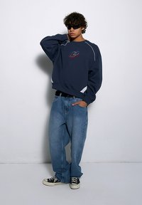 Granatowy sweter z logo, białe akcenty, luźny krój. Jasnoniebieskie dżinsy, oversize, proste nogawki. Czarne trampki z białą podeszwą.