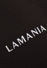 Balta siuvinėjimo "LAMANIA" tekstas juodame tekstūruotame fone, rodomas kampu.