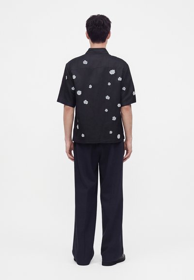 Homme de dos portant une chemise noire à manches courtes avec des imprimés floraux blancs et un pantalon ample foncé sur un fond blanc uni.