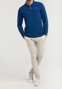 Långärmad blå pikétröja med krage, med en liten logotyp. Pairad med ljusbeiga byxor och vita sneakers. Enkelt design, avslappnad passform.