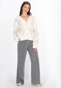 Cremfarbener Cardigan mit floralem Stickereimuster, V-Ausschnitt und Knopfverschluss. Kombiniert mit grauen, weiter geschnittenen Hosen und hellen Schuhen. Glatte Textur.