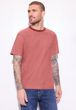 Mężczyzna z krótkimi włosami i tatuażami na ramionach, ubrany w czerwoną koszulkę w paski i niebieskie dżinsy, patrzący w lewo na jednolitym białym tle.