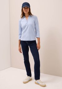 Kobieta w jasnoniebieskiej koszulce polo z długim rękawem, ciemnoniebieskich dżinsach, białych trampkach z niebieskimi akcentami i dżinsowej czapce z daszkiem stoi przy beżowej ścianie.