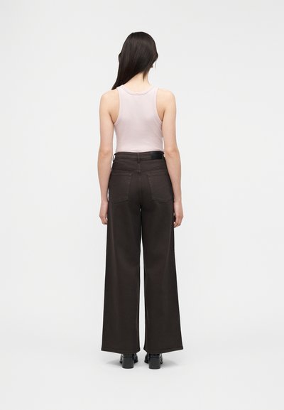 Γυναίκα με μακριά σκούρα μαλλιά, που φορεί αμάνικο ανοιχτό ροζ τοπ, καφέ ψηλόμεσο παντελόνι με φαρδιές παντελόνες και μαύρες γόβες, στέκεται γυρισμένη προς τα πίσω.