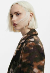 Brun og grøn mønstret frakke med en blød, fuzzy tekstur. Har en klassisk krave og en enkelt knaplukning foran.