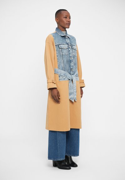 Robe en denim et coton avec un haut en denim bleu et un bas en jaune moutarde. Elle dispose d'une ceinture à la taille et d'un col, associée à un pantalon en denim à jambes larges.