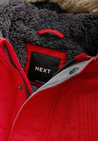 Röd jacka med grå innerfoder, med en strukturerad, fluffig krage, dragkedjor och en svart etikett med texten "NEXT."