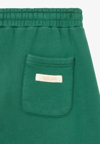 Pantalons de survêtement verts avec une taille élastique, comportant une poche arrière et une étiquette cousue dans la poche. Texture lisse et design décontracté.