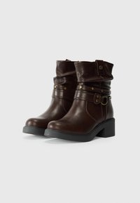 Brown mid-calf leather boots with decorative straps and metallic studs, stacked heels, and rounded toes on a white background.