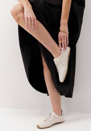 Persona con vestido negro levanta una pierna, mostrando una zapatilla beige con cordones, logo y joyas doradas en la mano sobre un fondo blanco.
