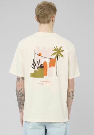Personne portant un t-shirt beige avec un design abstrait coloré représentant des marches, des plantes, un palmier et un tissu orange au dos.